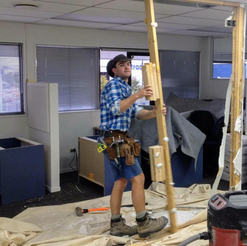 Matthew Horncastle wearing a tool belt and holding timber inside a room being renovated